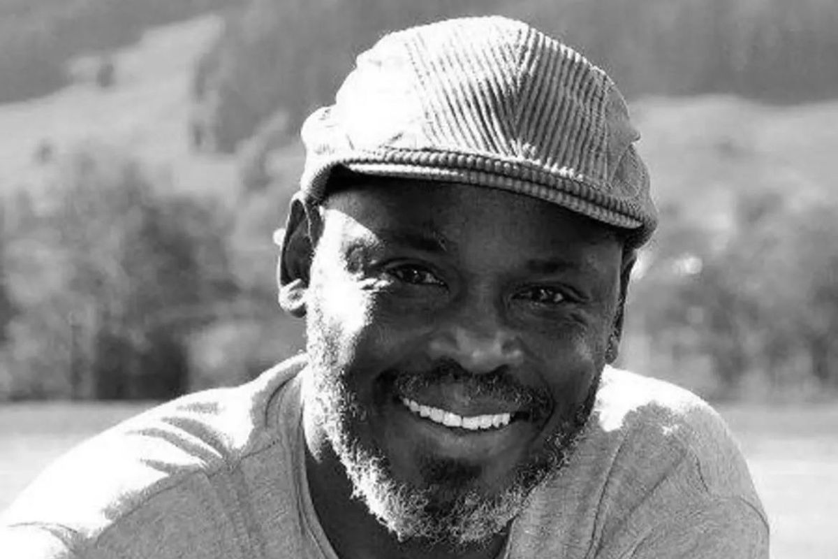 Black and white headshot of Abdelaziz Baraka Sakin, smiling into the camera and wearing a cap. 
