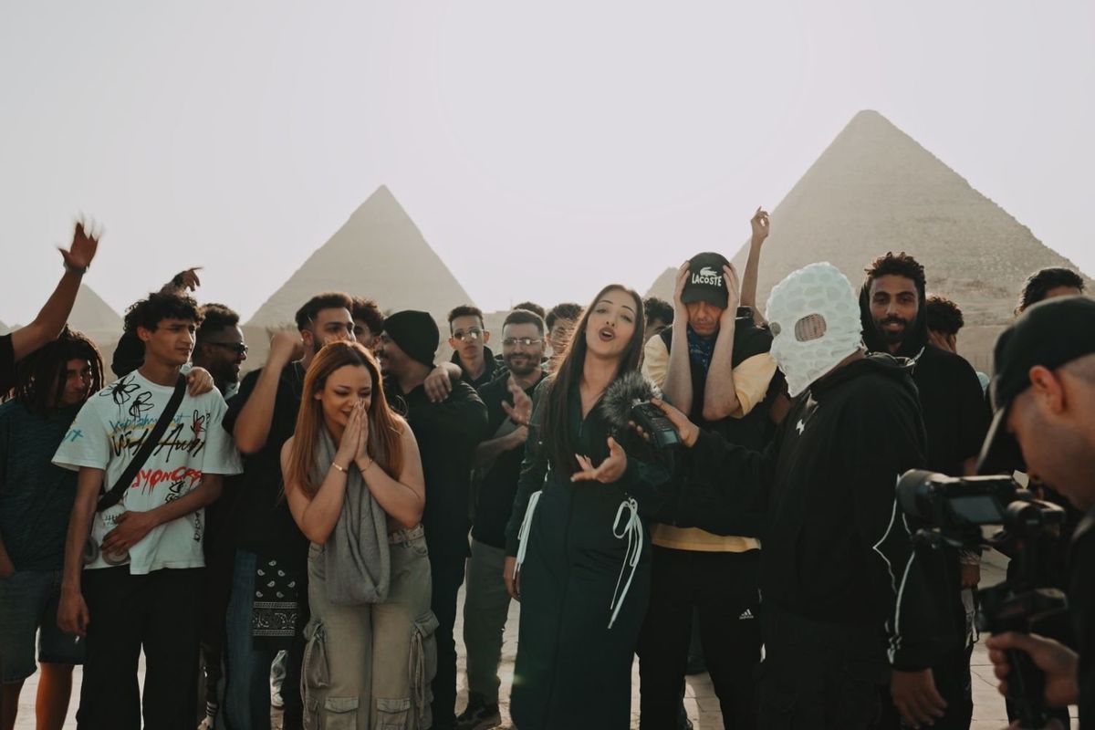 Egyptian rappers gathered in front of the Great Pyramids of Giza, one masked man is holding a microphone to a young woman rapping in the middle of the group. 